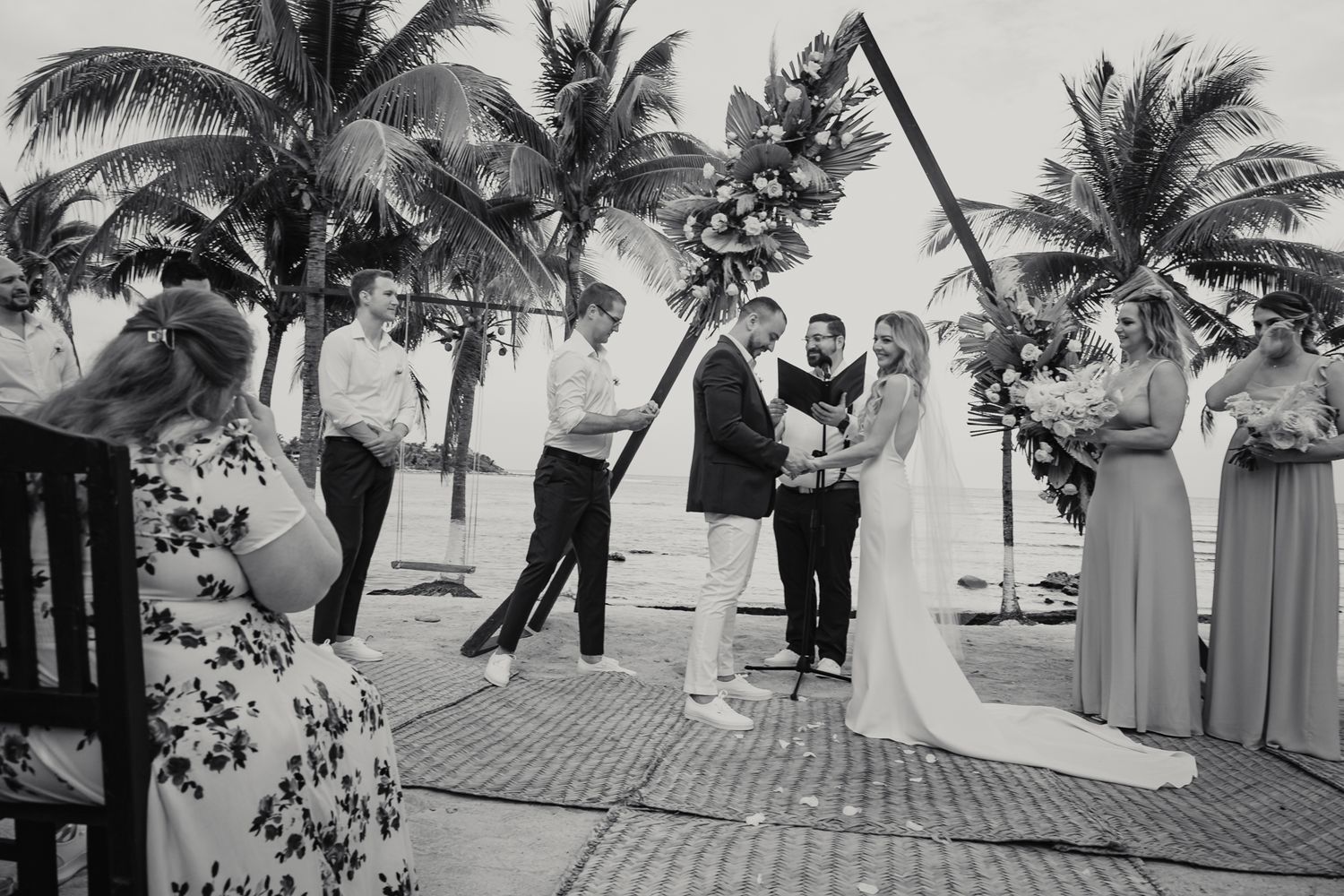 Couple exchanges vows at a tropical beach wedding ceremony under a triangular floral arch.