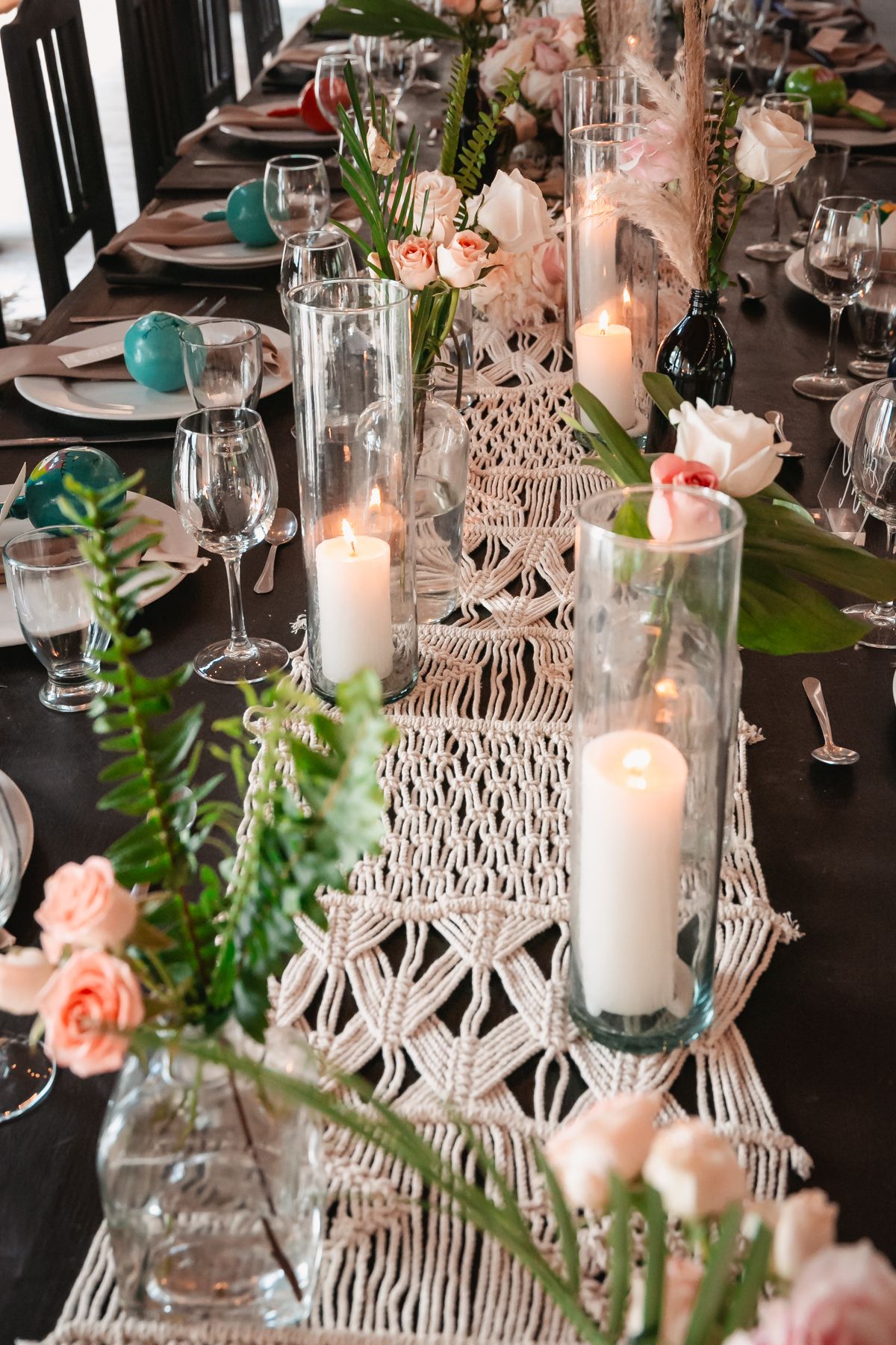Elegant wedding reception table with macrame runner, white candles, pink roses, and glassware.