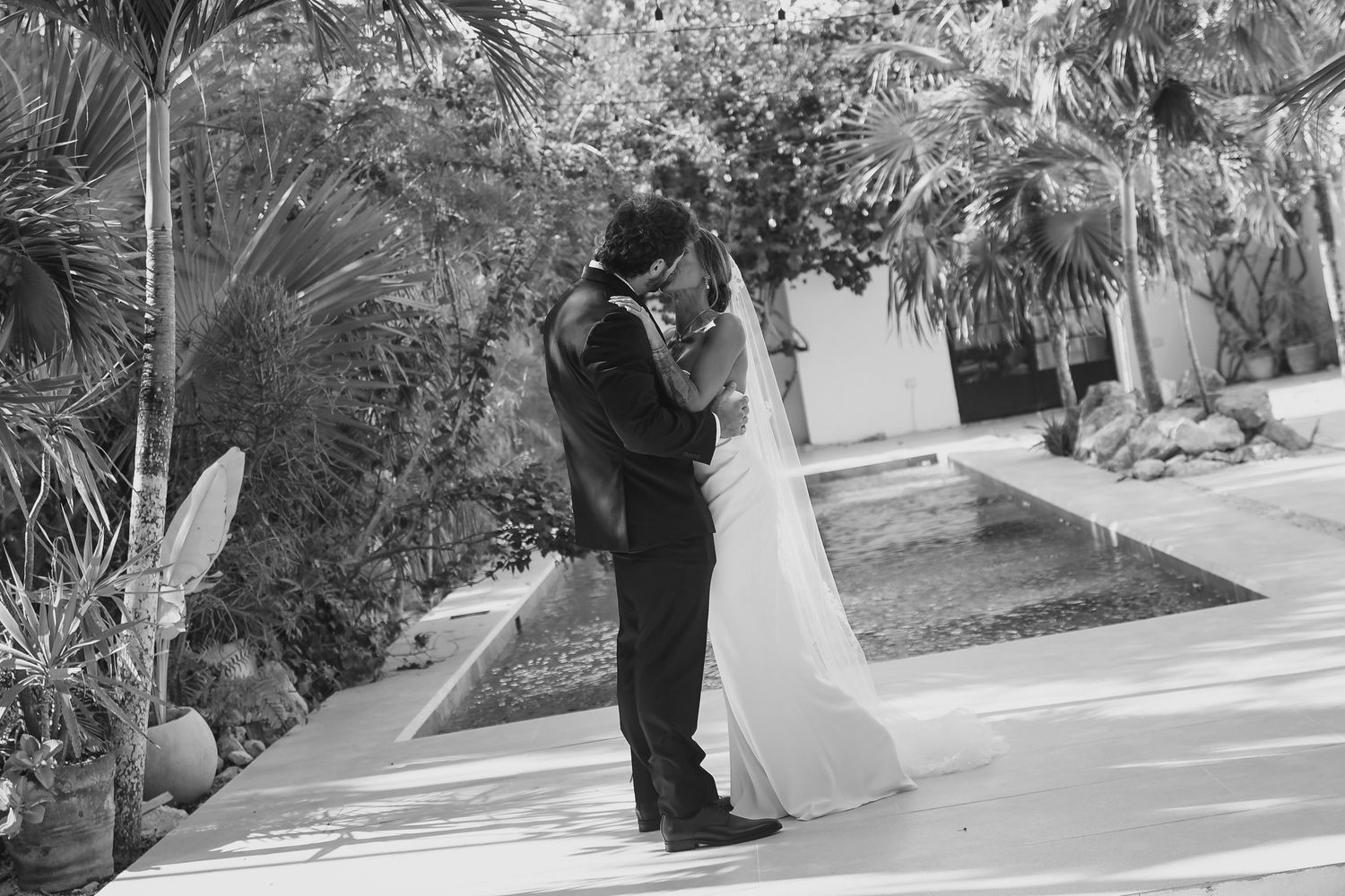 Black and white wedding photo of bride and groom embracing on a garden path with tropical foliage.