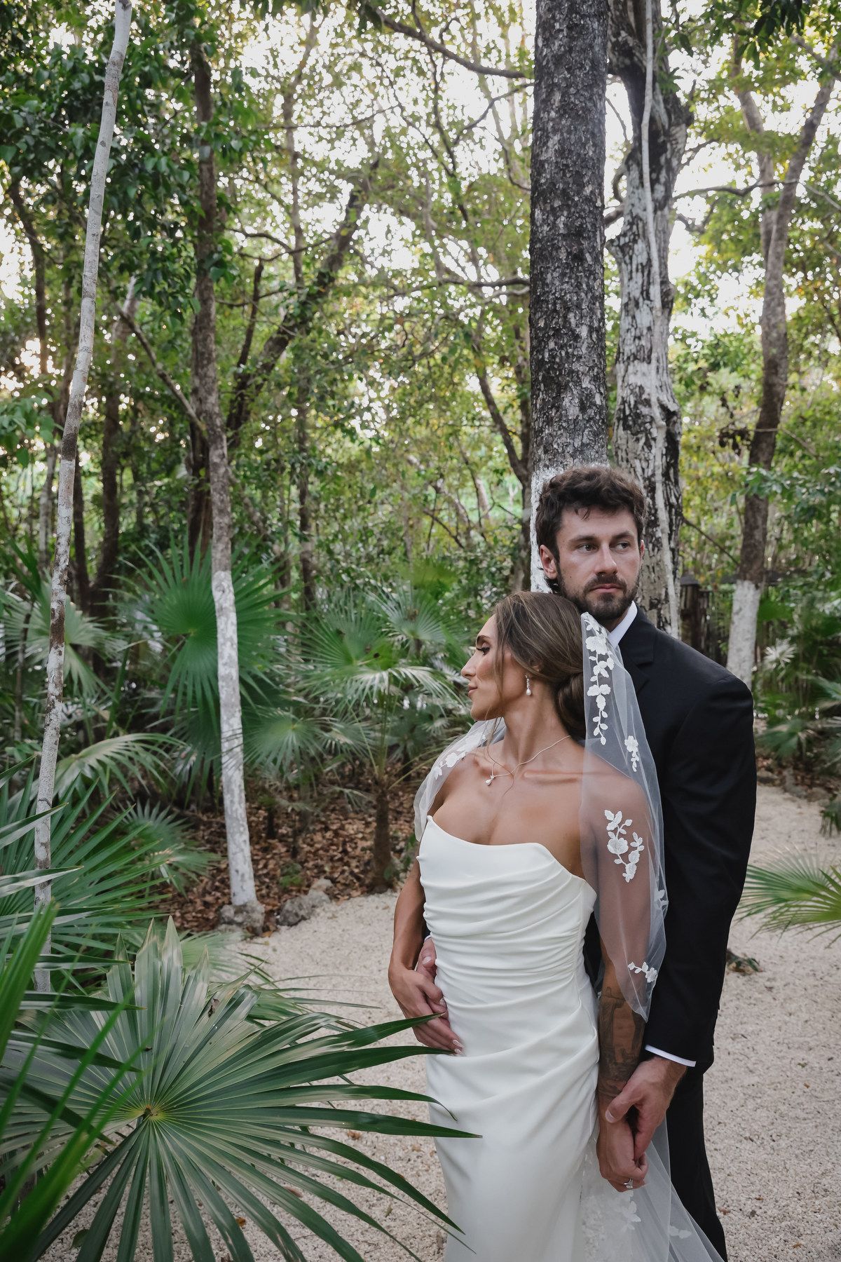 Bride and groom pose romantically in a lush tropical forest, she in a white gown with floral veil, he in a black tuxedo.