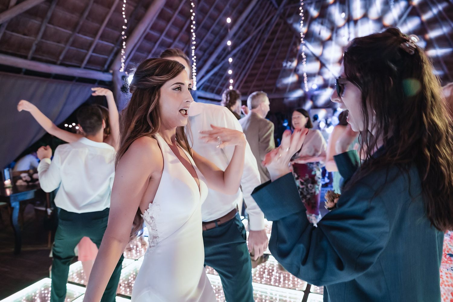Bride in white gown dancing with guest in teal dress on mirrored wedding dance floor.