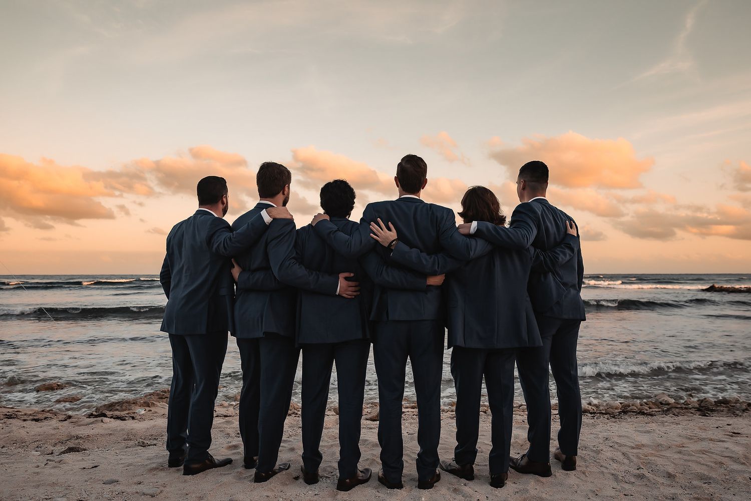 Six men in dark suits stand arm in arm on a beach at sunset, facing the ocean.