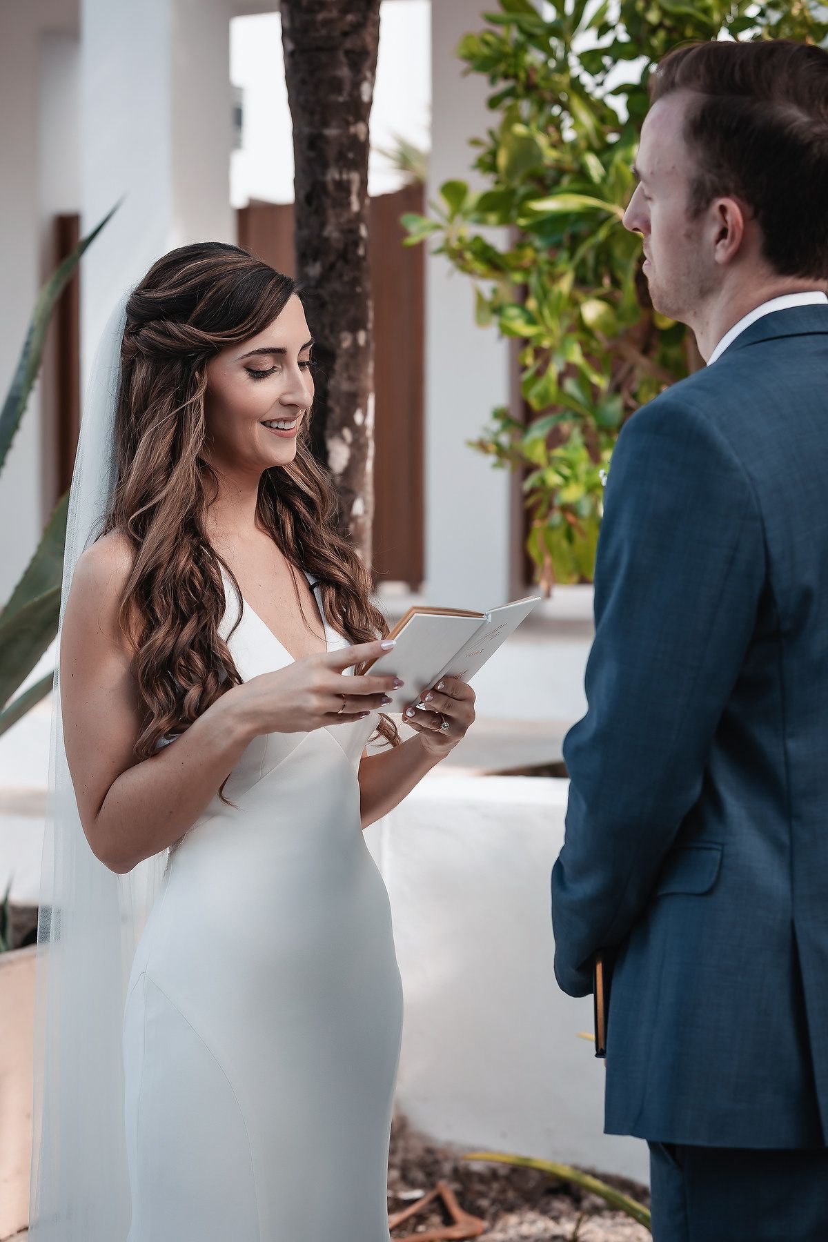 Bride in white gown reads wedding vows from a card to groom in blue suit during outdoor ceremony.