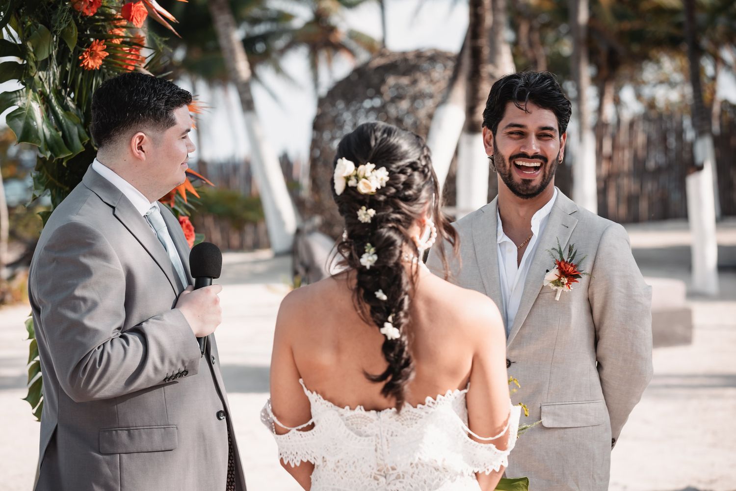 Groom in tan suit smiles at bride during outdoor beach wedding ceremony with officiant.