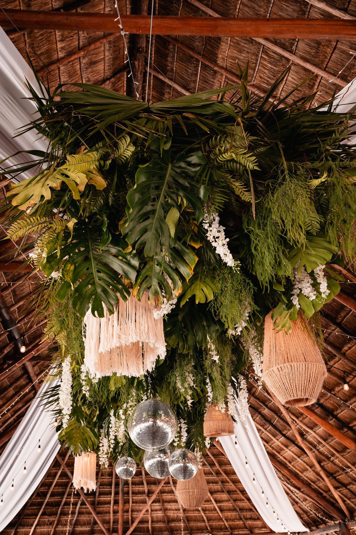 Tropical wedding ceiling installation with monstera leaves, ferns, disco balls, and woven lanterns in a rustic thatched venue.