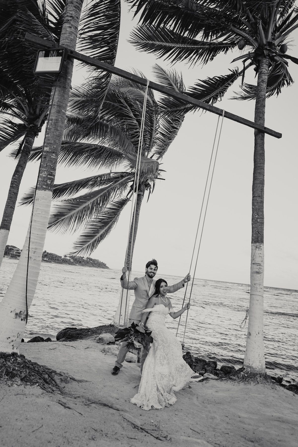 Bride and groom sharing romantic moment on rope swing beneath palm trees on tropical beach wedding day.