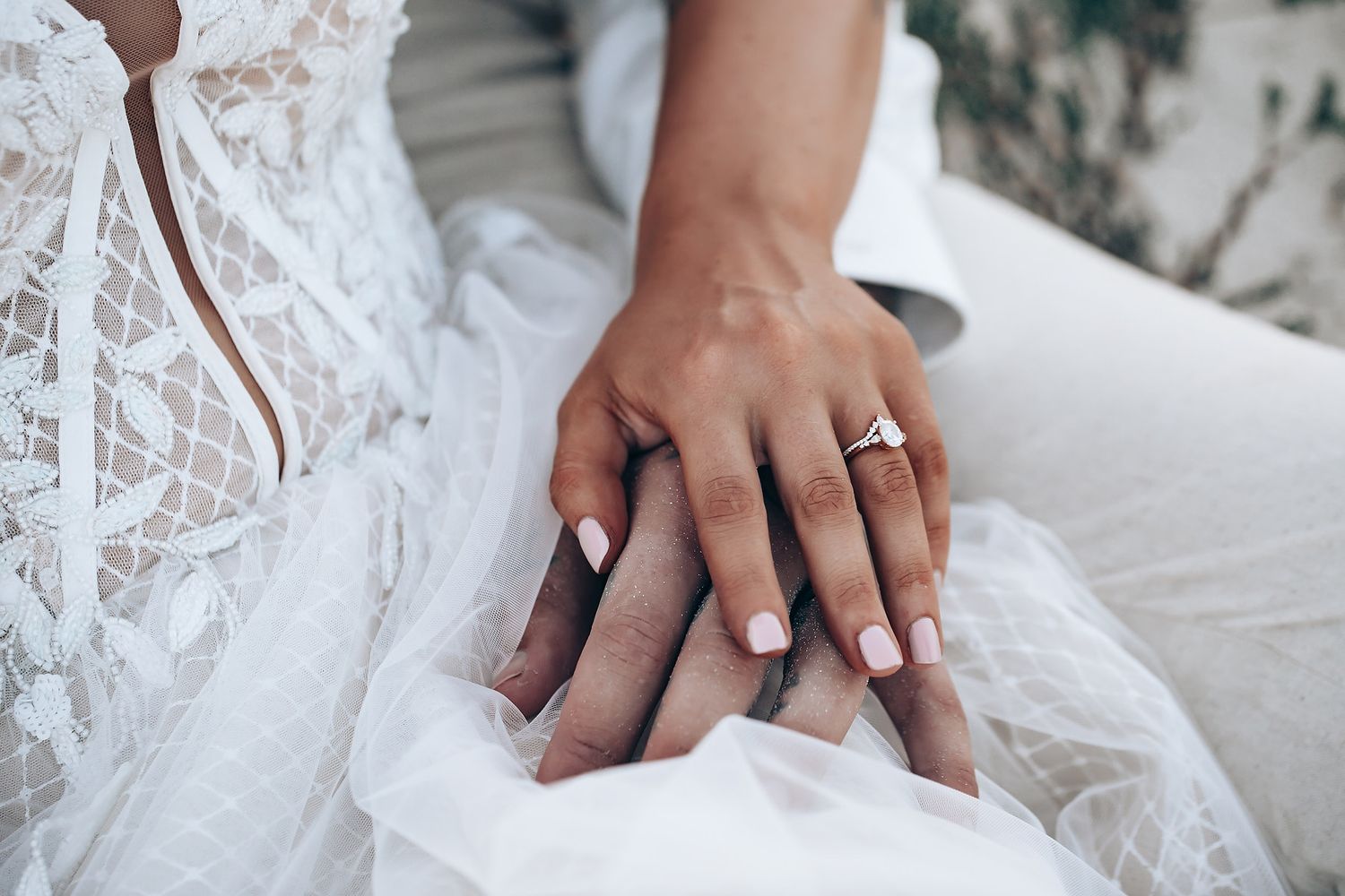 Close-up detail of delicate white lace wedding dress fabric and hands with rings.