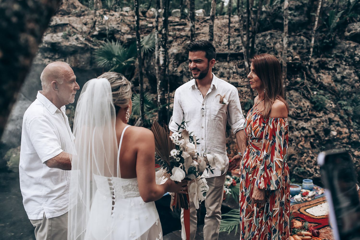 Wedding guests gather in a rustic outdoor setting with stone walls and natural greenery.