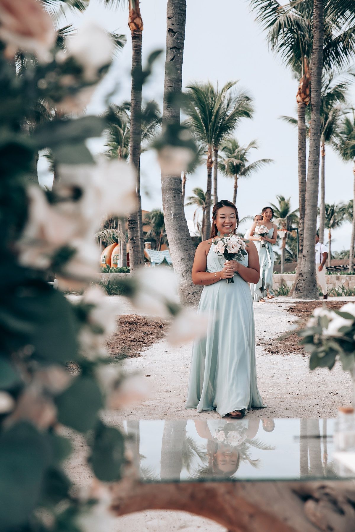 A bridesmaid in a mint green dress holds a bouquet while standing on a tropical beach path lined with palm trees.