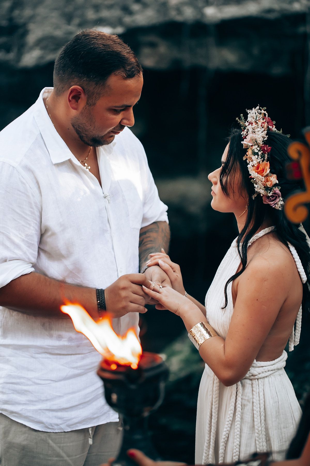 A romantic ceremony with white attire and floral crown takes place by firelight in a tropical setting.