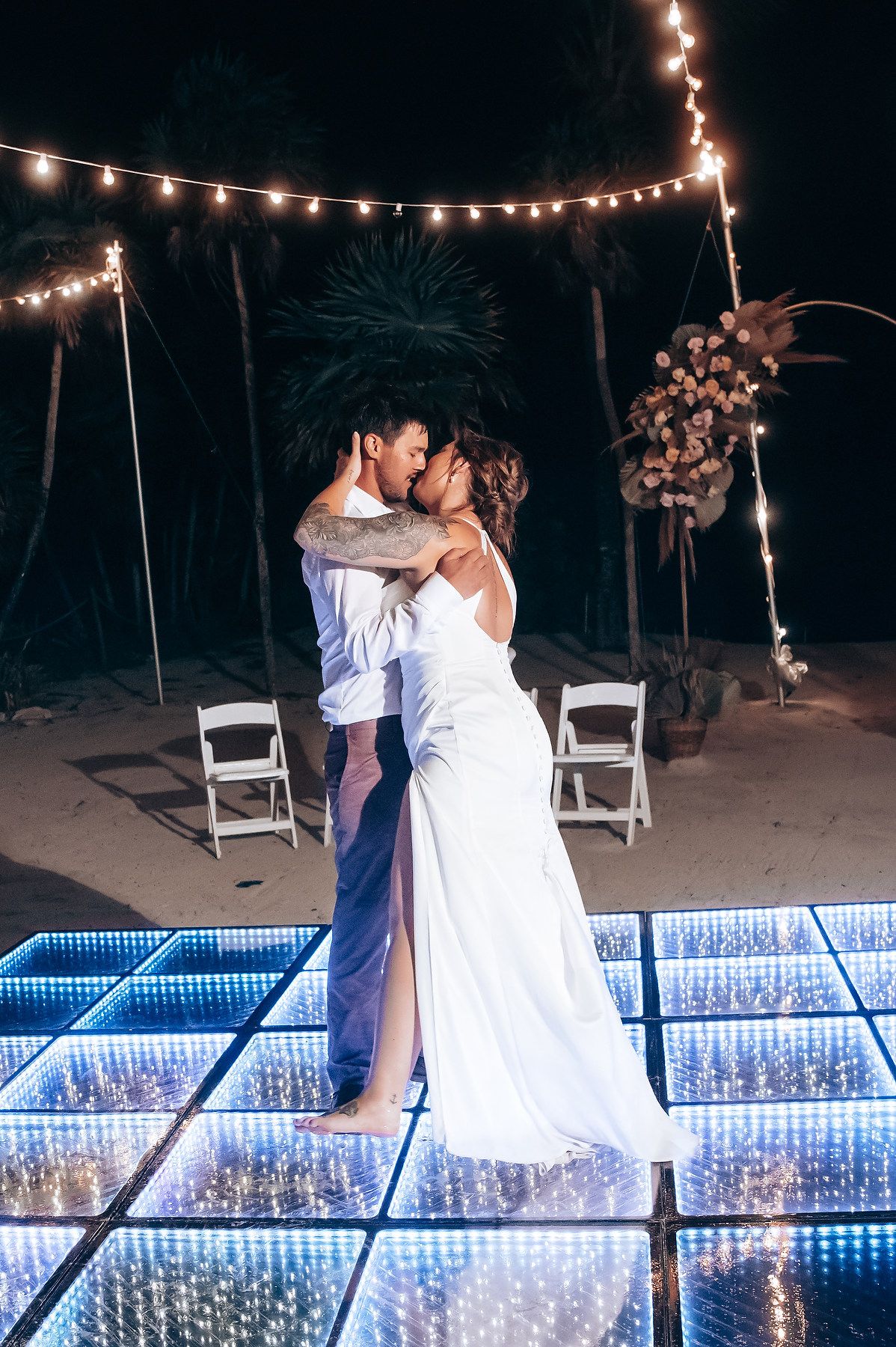 A romantic evening dance under string lights by an illuminated pool with palm trees in the background.