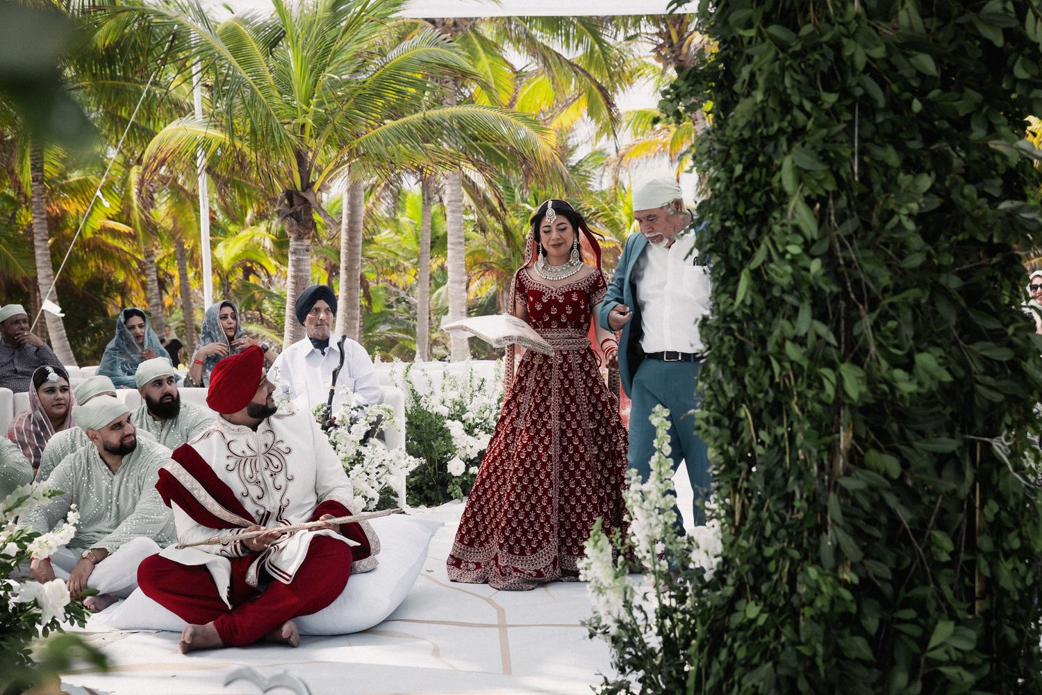 Bride in red lehenga walks during Sikh wedding ceremony under floral mandap with palm trees.