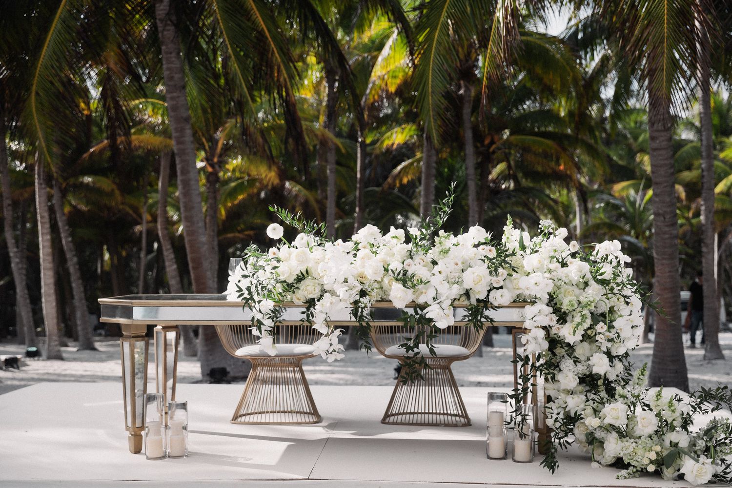 Elegant outdoor wedding reception setup with white floral arrangements on rattan stands amid tropical palm trees.