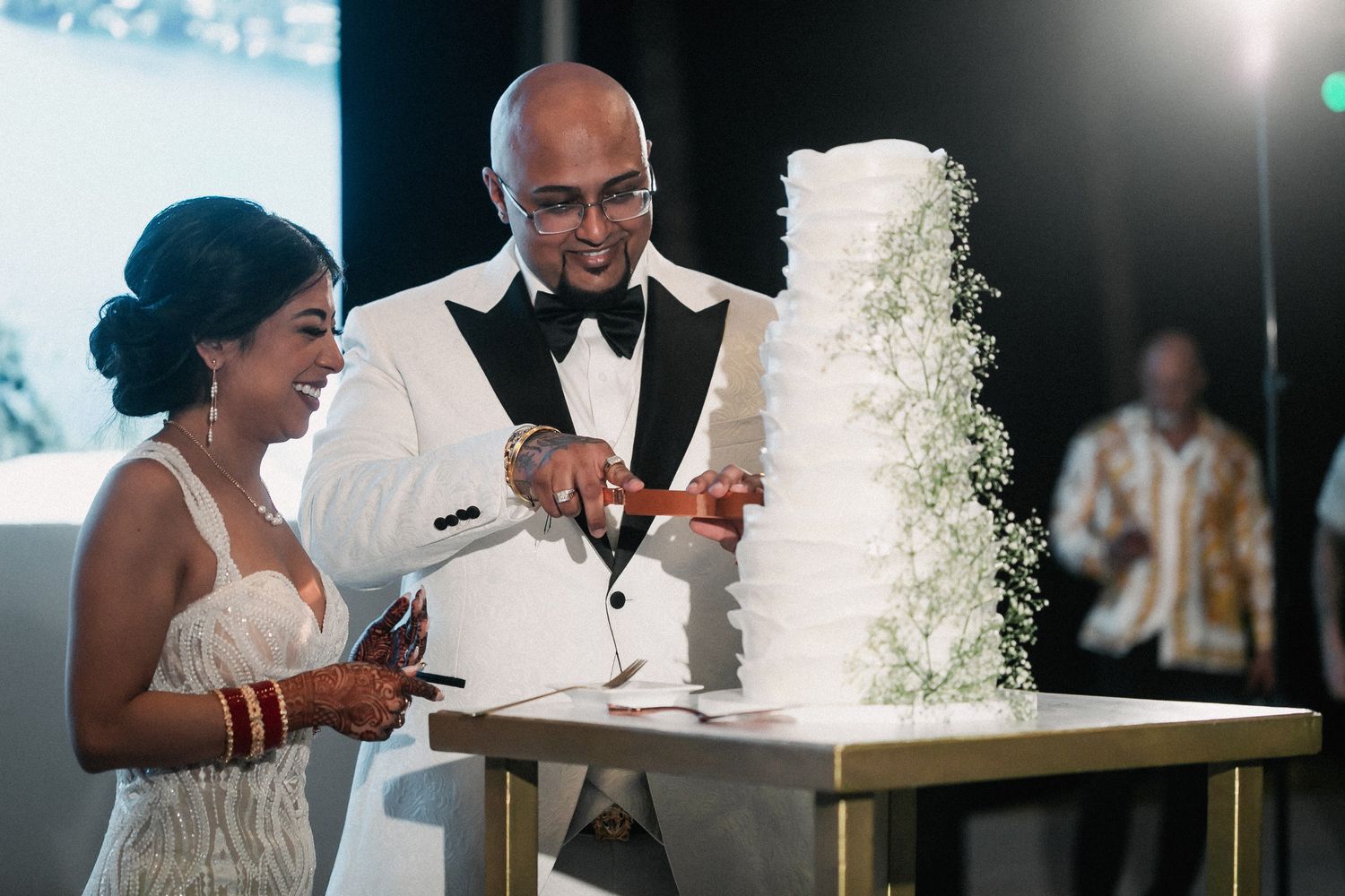 Indian bride and groom cut tall white wedding cake adorned with baby's breath at reception.