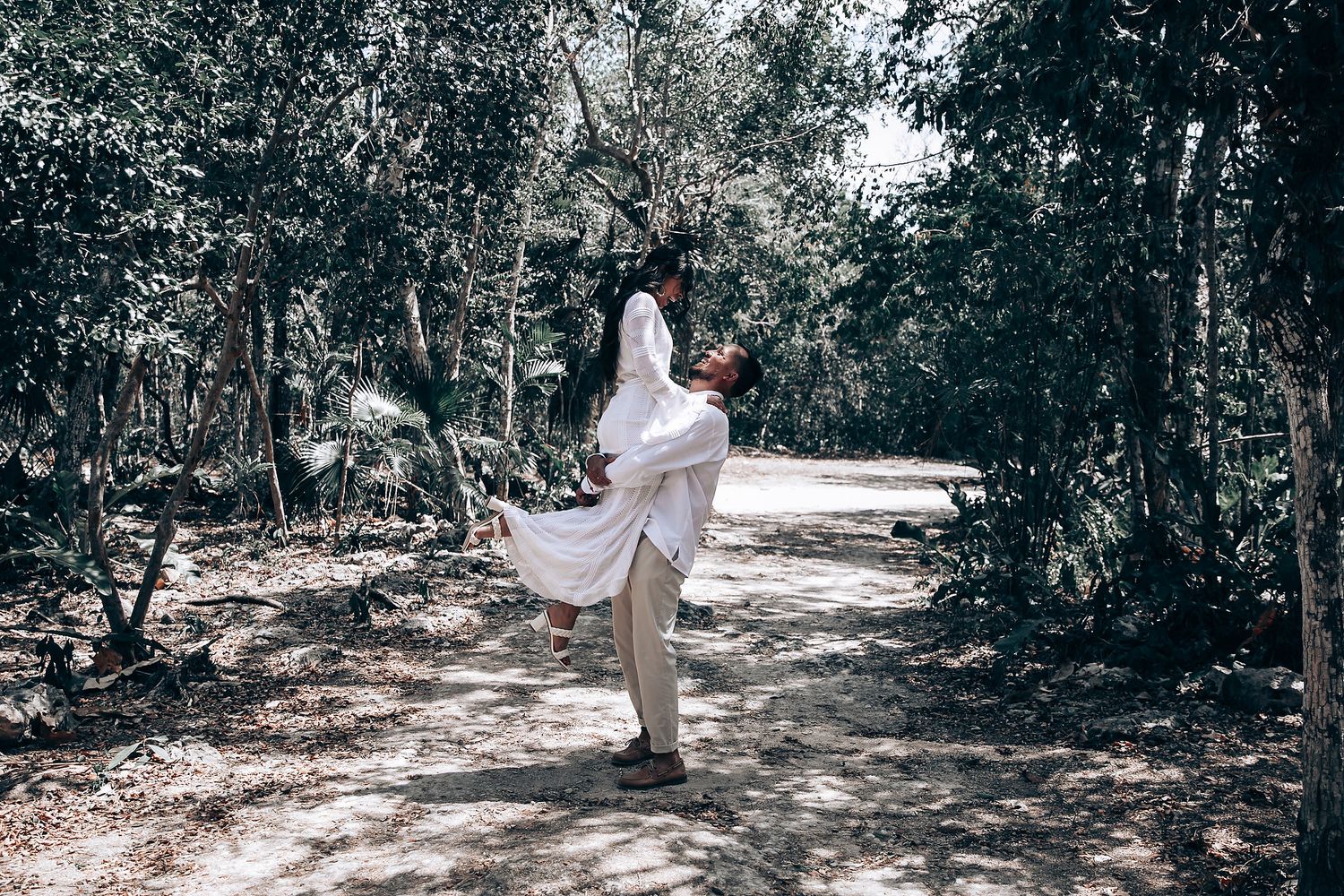 A couple in matching white attire walk together through a serene forest setting.