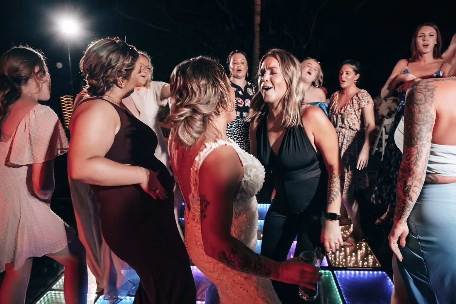 Bride and guests dance joyfully together on a colorfully lit dance floor at wedding reception.