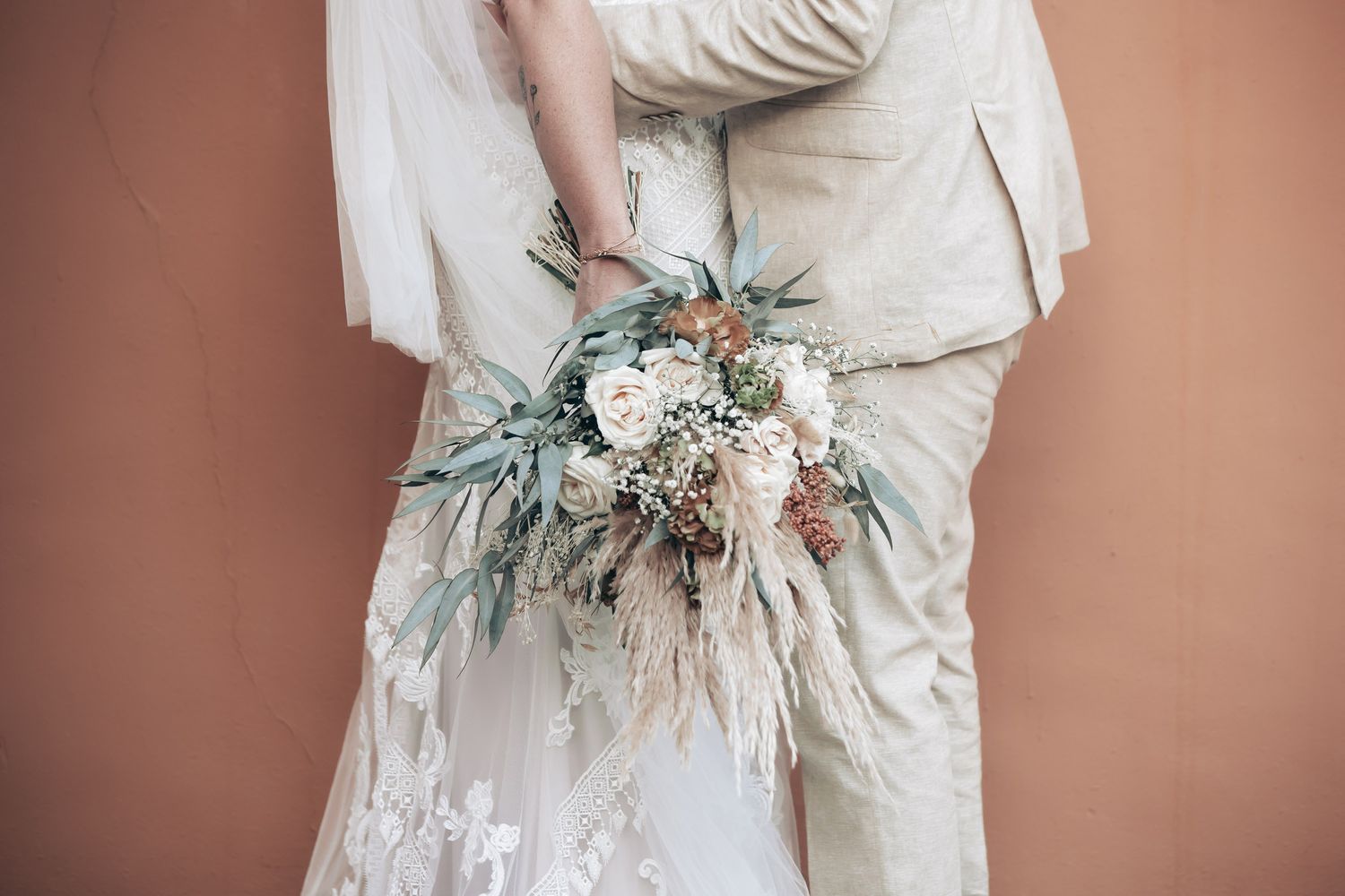 Bride and groom embracing while holding rustic boho wedding bouquet against warm wall.