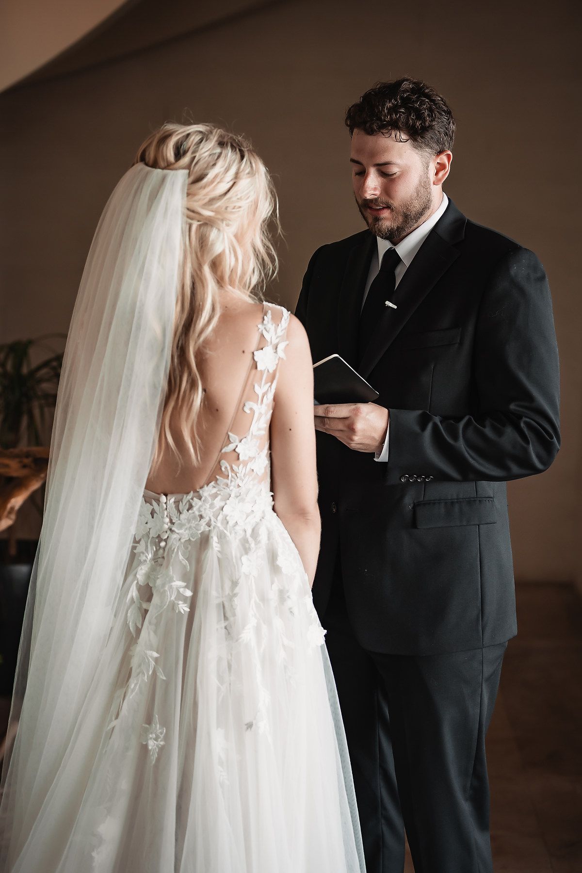 Bride in lace wedding gown with flowing veil during first look with groom in black suit.