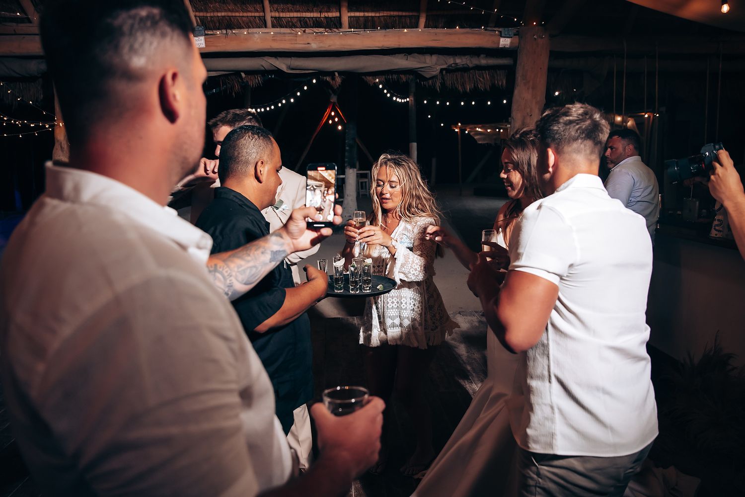 A group of people dancing and celebrating at a dimly lit wedding reception venue with string lights overhead.