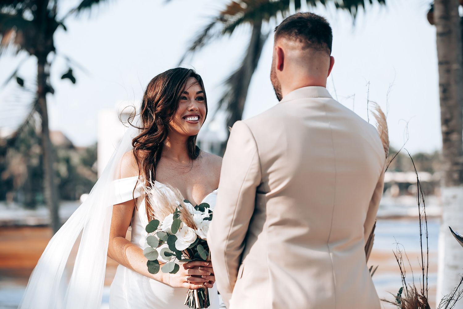 Beachfront wedding ceremony with elegant white decor and palm trees swaying in the background.