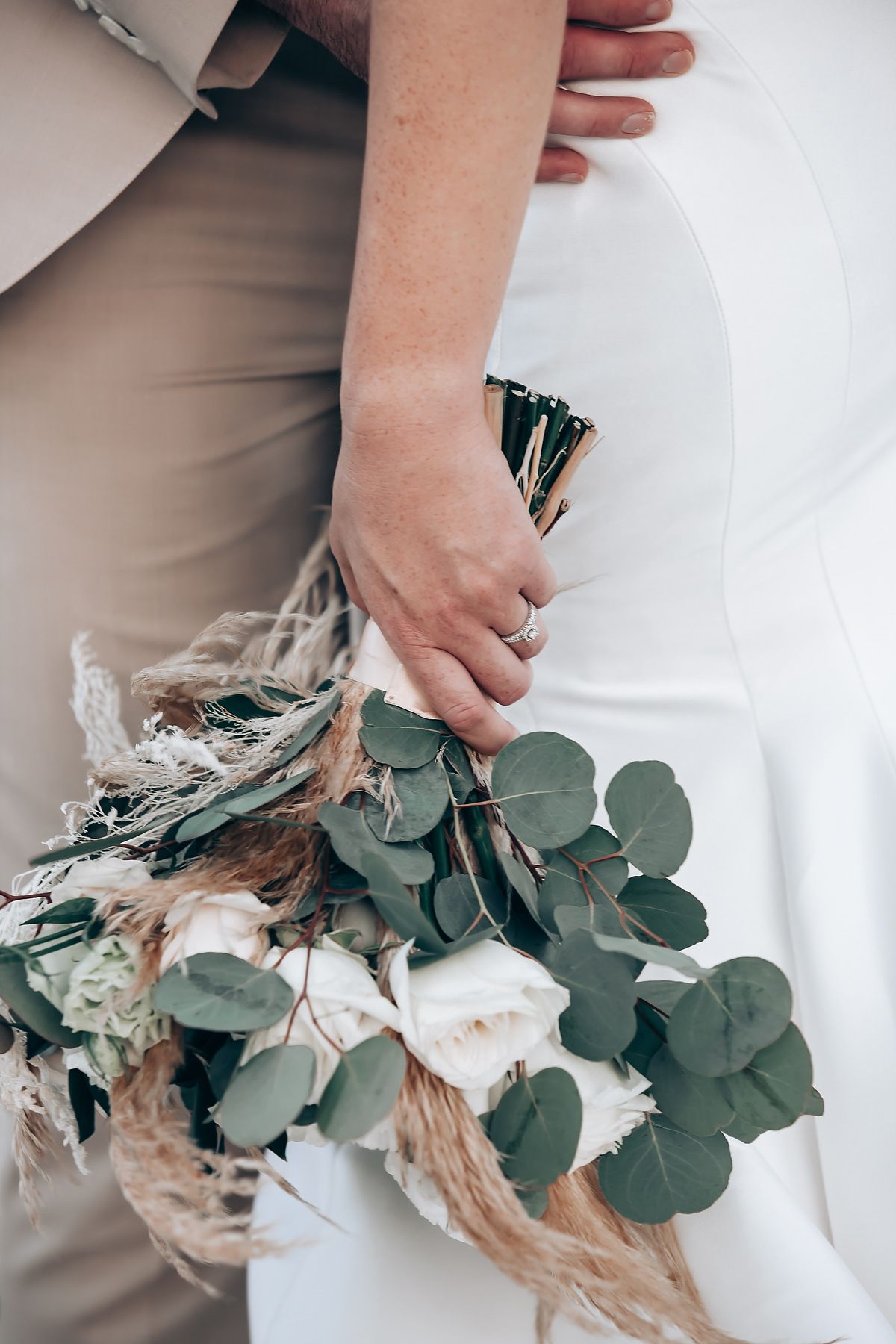 Close-up view of hands holding a rustic bouquet with eucalyptus leaves and dried natural elements against white background.