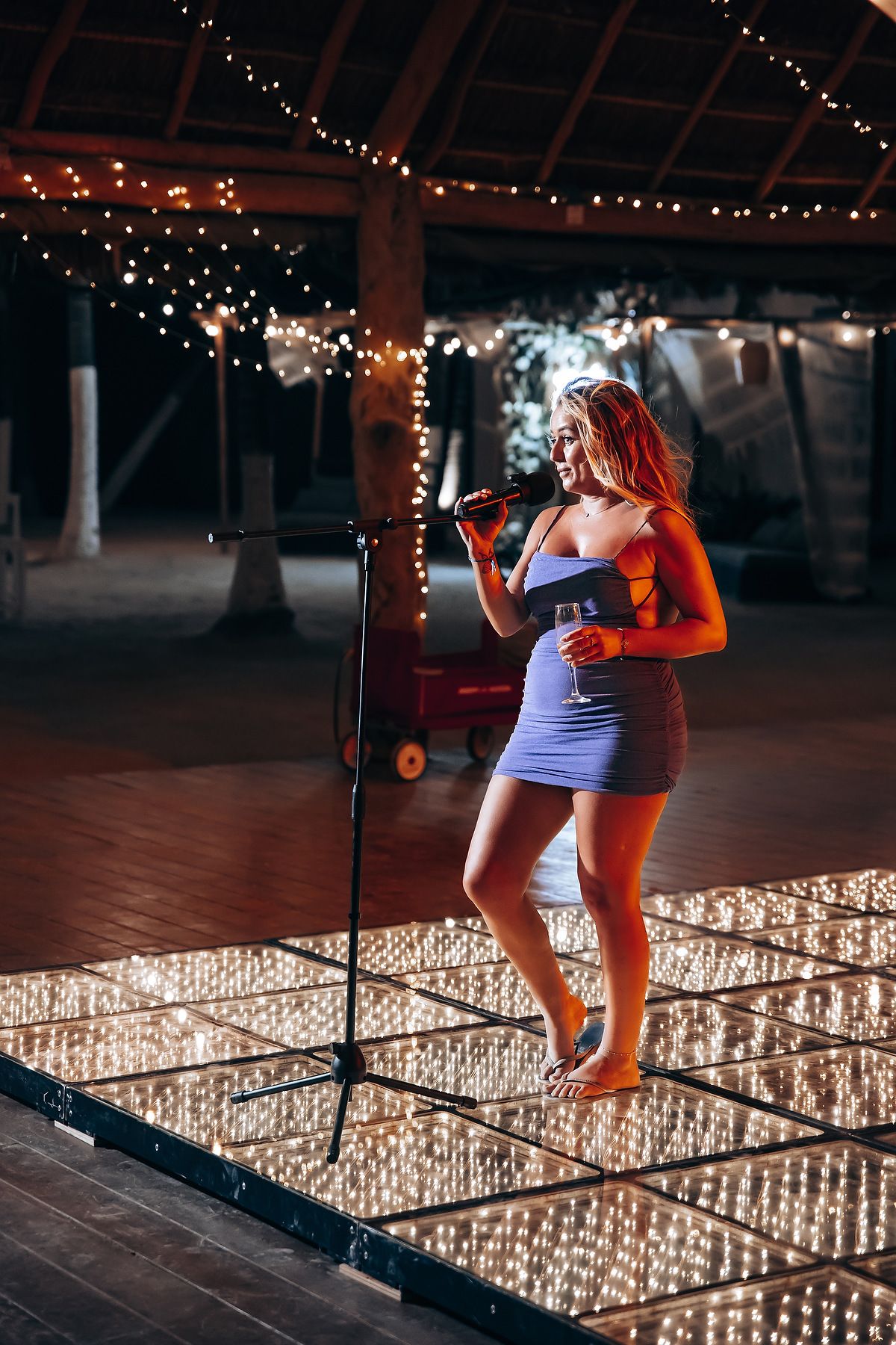 A figure stands on an illuminated dance floor at night under string lights in a short dress holding a microphone.
