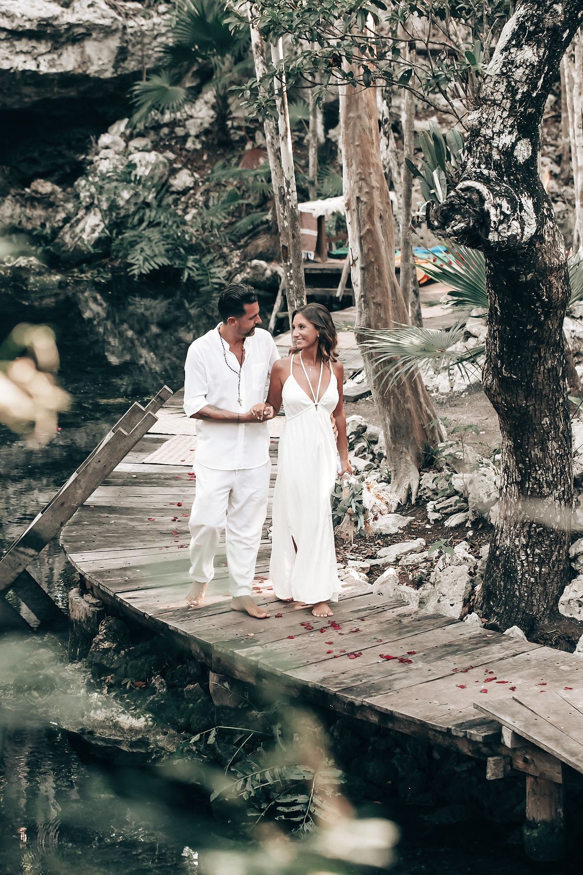 A couple in white clothing stands on a wooden bridge in a lush, natural setting.