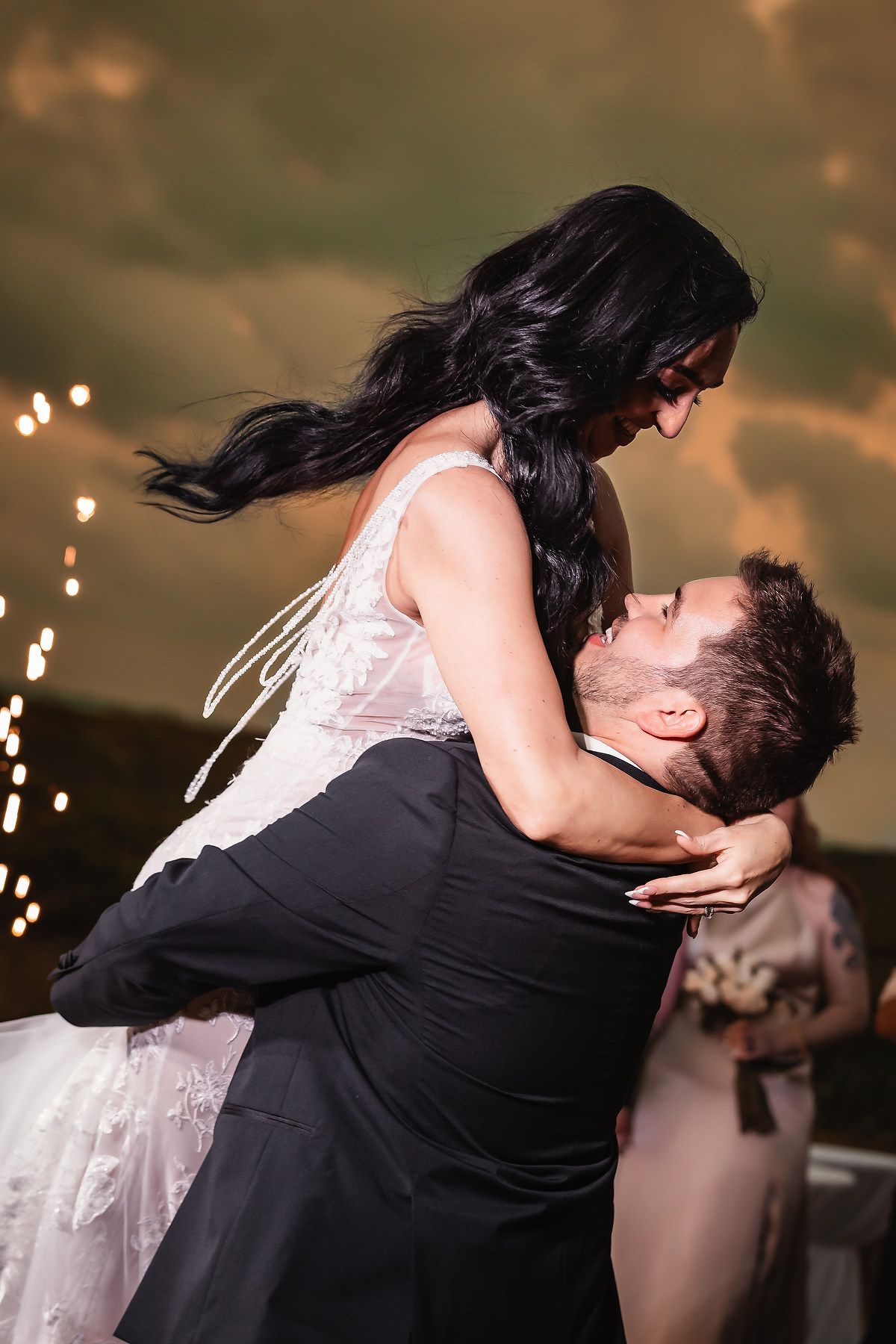 Groom lifting bride in white dress during romantic first dance with sparkling lights at wedding reception.