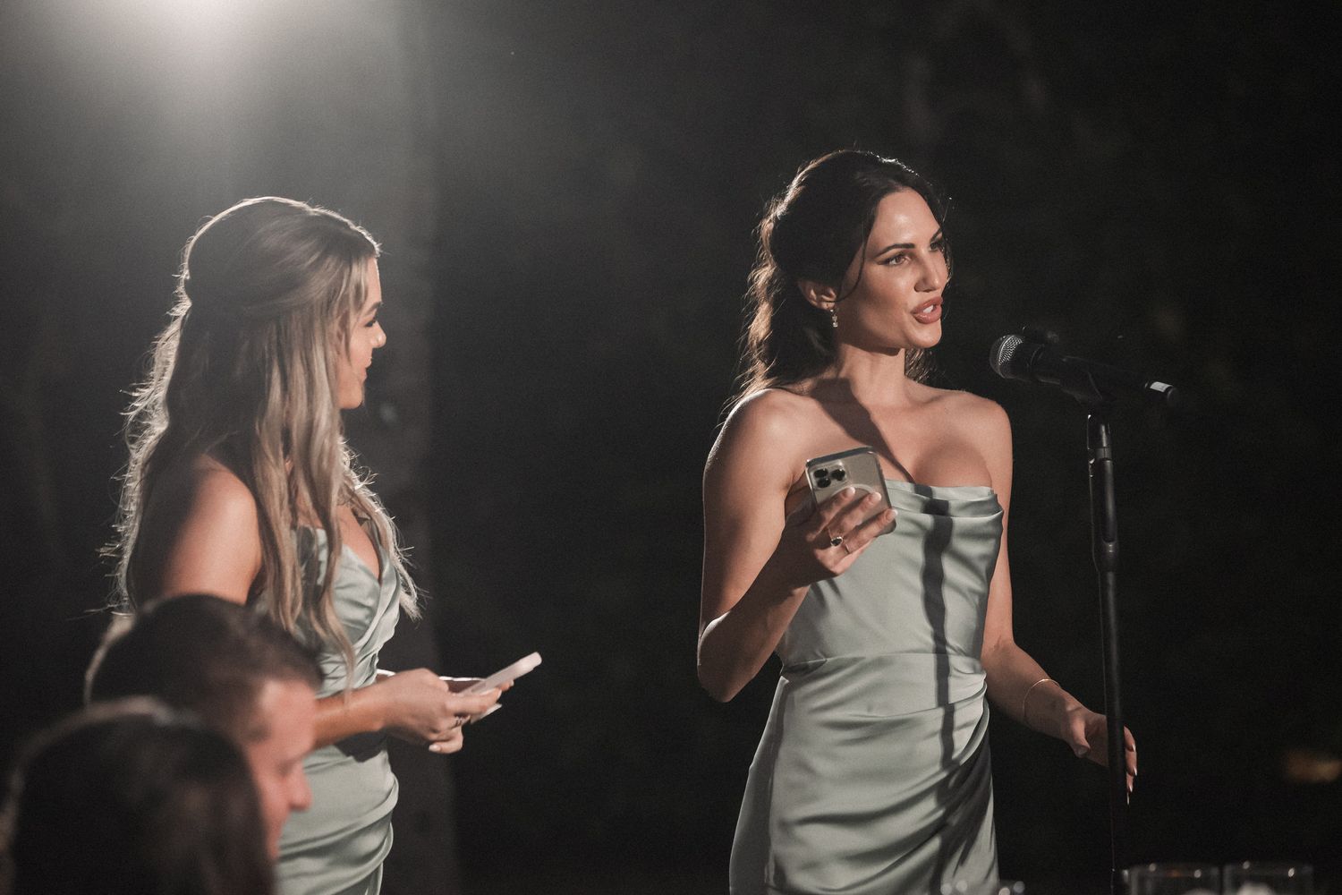 Two bridesmaids in sage green dresses giving speeches at microphone during elegant wedding reception.