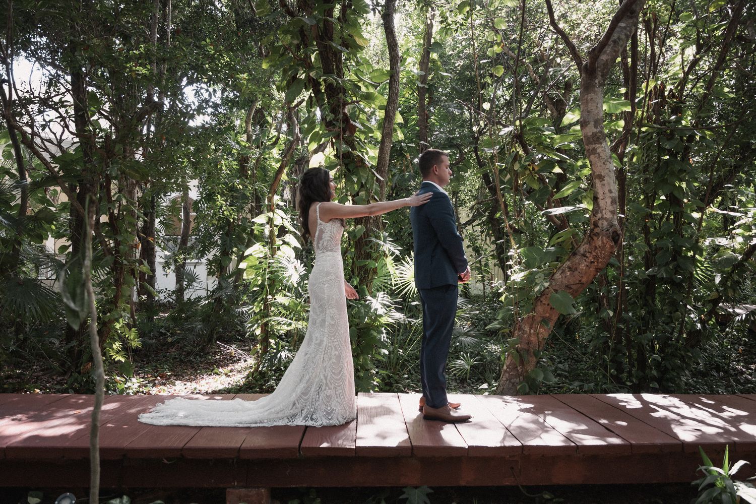 Bride and groom share first look moment on wooden deck surrounded by lush tropical forest trees.