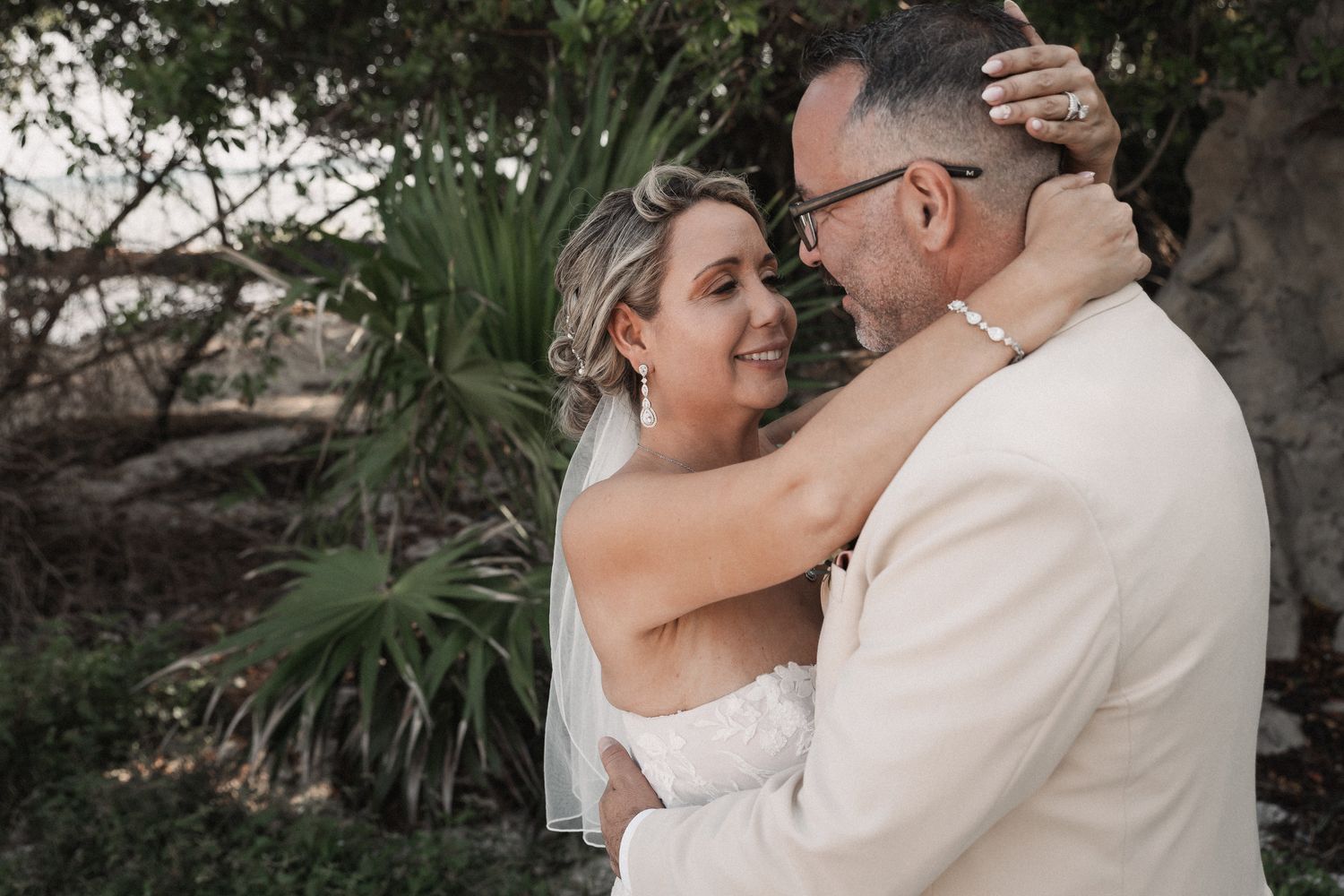 Smiling bride in white gown and veil embracing groom in beige suit amid tropical greenery.