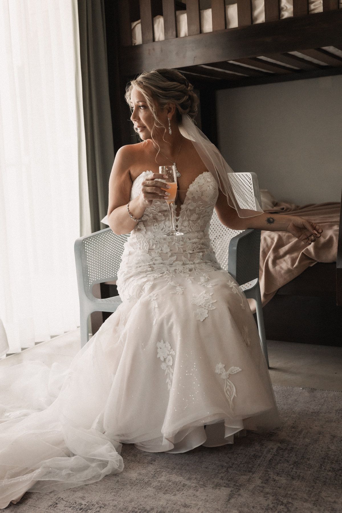Bride in strapless lace mermaid wedding gown holding champagne flute while seated near window with veil.