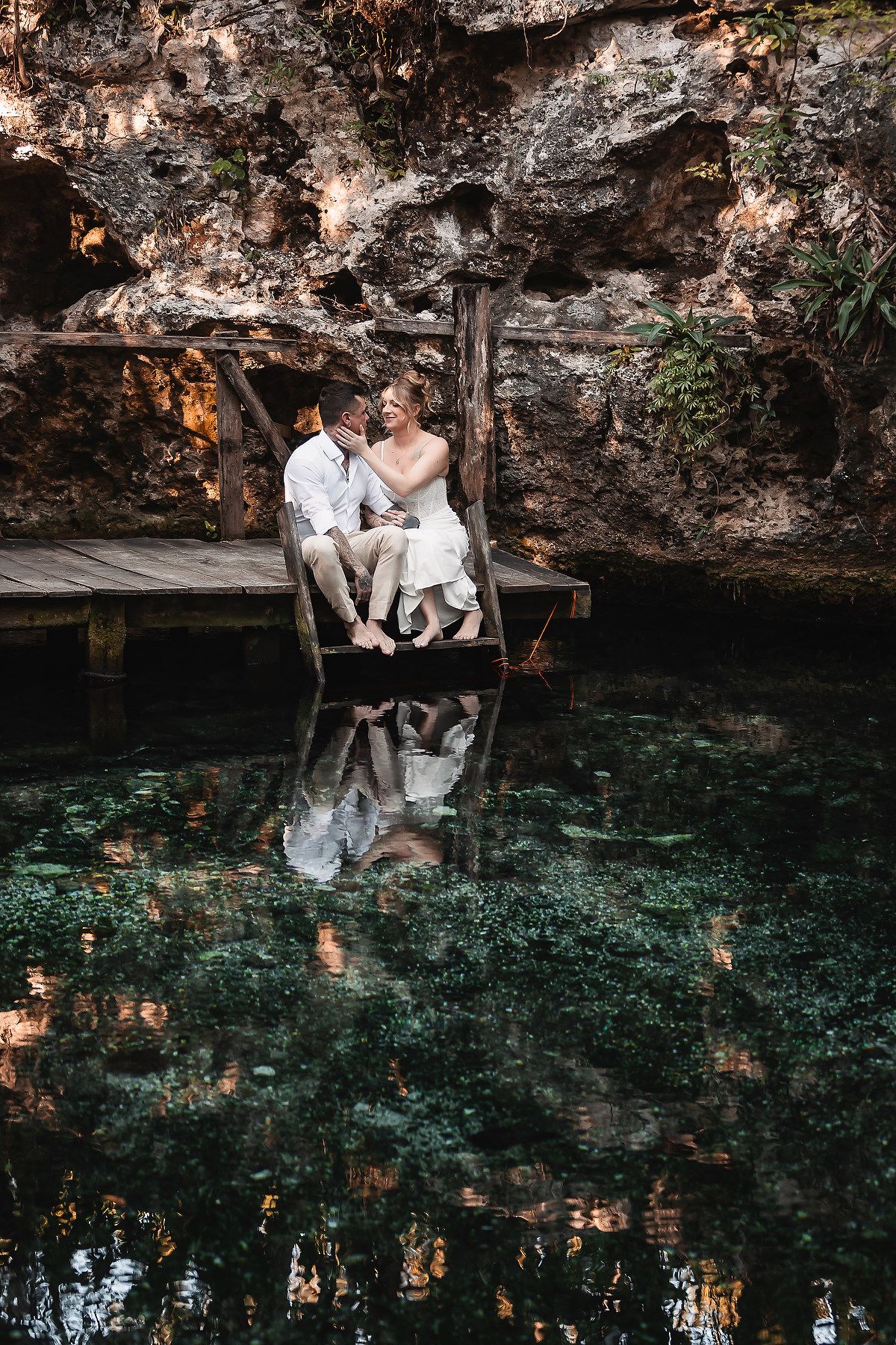 Couple sitting on a wooden dock over a serene cenote, reflecting lush greenery and rocky walls in still water.