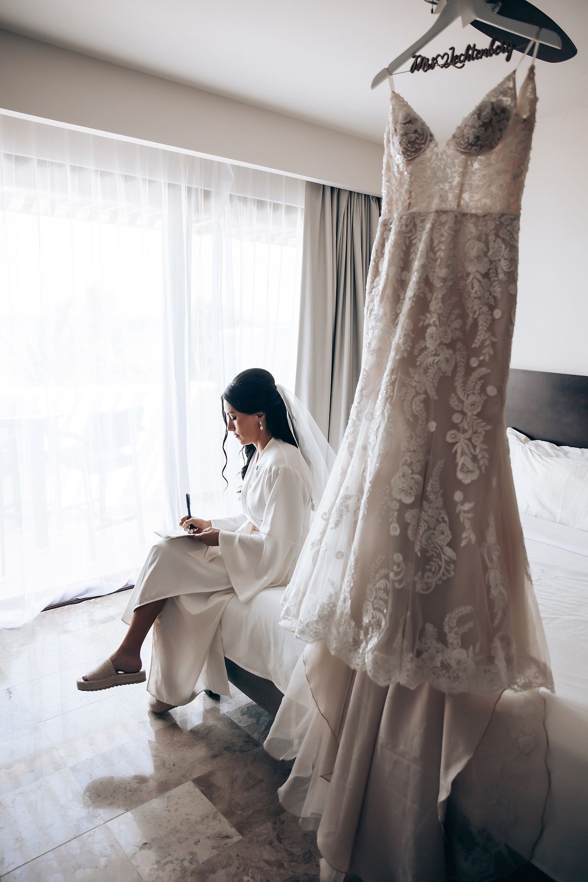 Elegant lace wedding dress hangs by window in bridal preparation room showing delicate details and flowing silhouette.