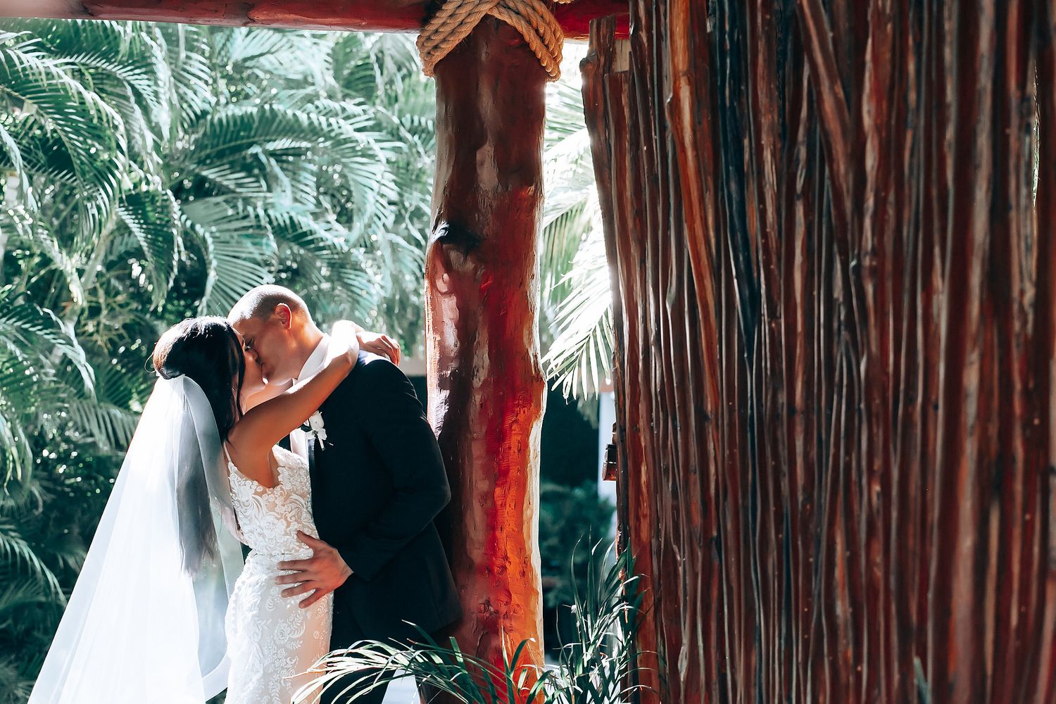 Wedding couple shares intimate moments under a rustic wooden gazebo surrounded by tropical palm trees.