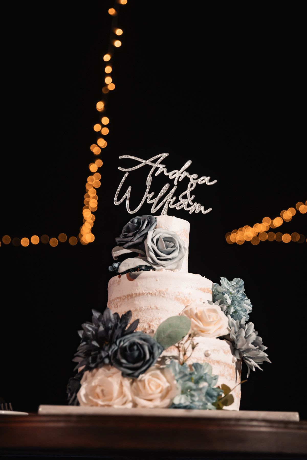 Three-tier white wedding cake with blue and cream roses, topped with Andrea & William cake topper, bokeh lights in background.