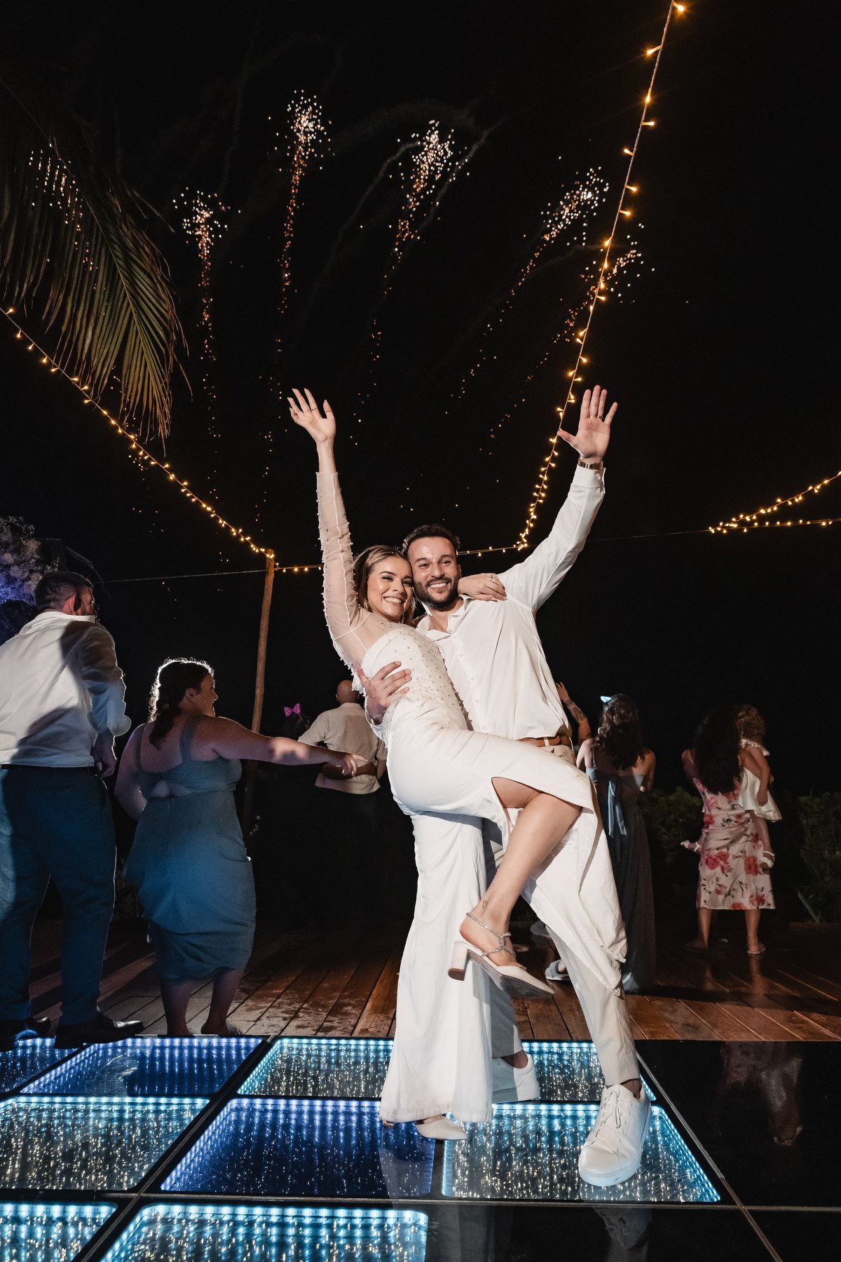 Joyful bride and groom dancing and celebrating on lit dance floor at night reception.