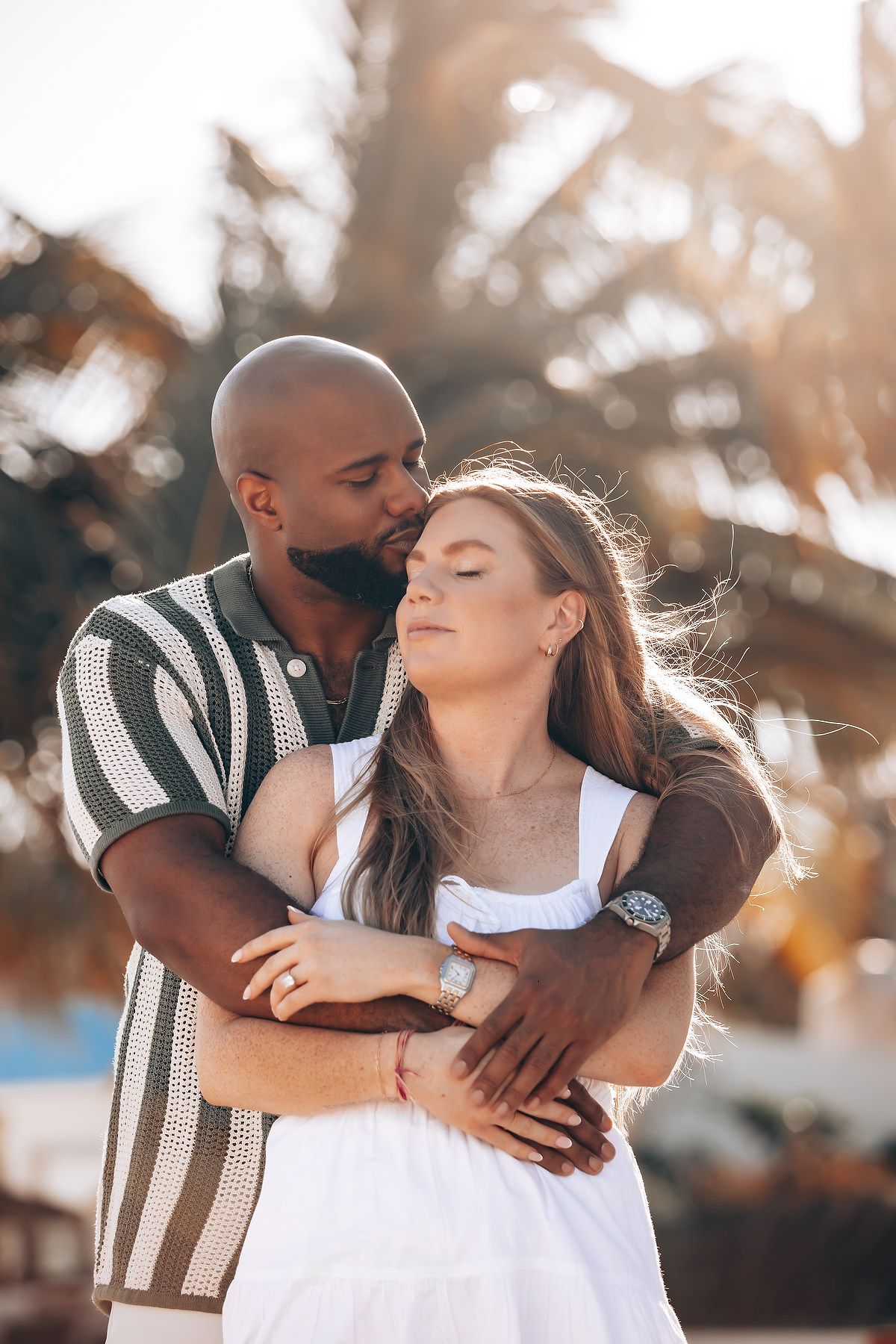 Man in striped shirt tenderly embracing woman in white dress at golden sunset near palm trees.