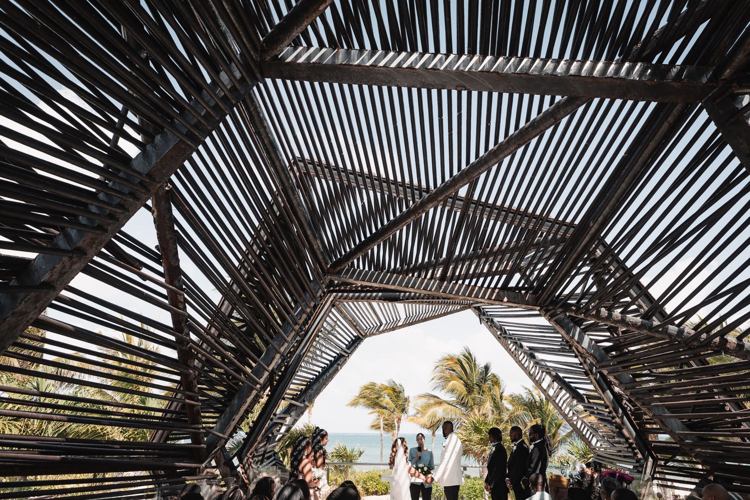 Wedding ceremony under a stunning geometric bamboo arch structure with ocean view backdrop.