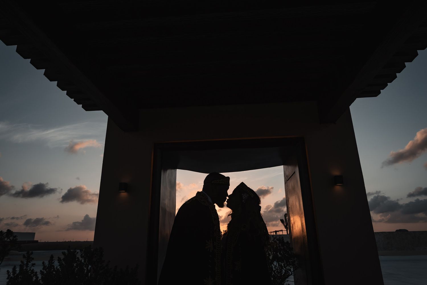 Silhouette of bride and groom sharing intimate moment framed by archway at sunset.