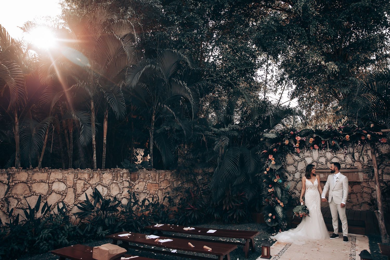 Sunlit outdoor wedding ceremony with guests seated in rows among lush tropical landscaping.