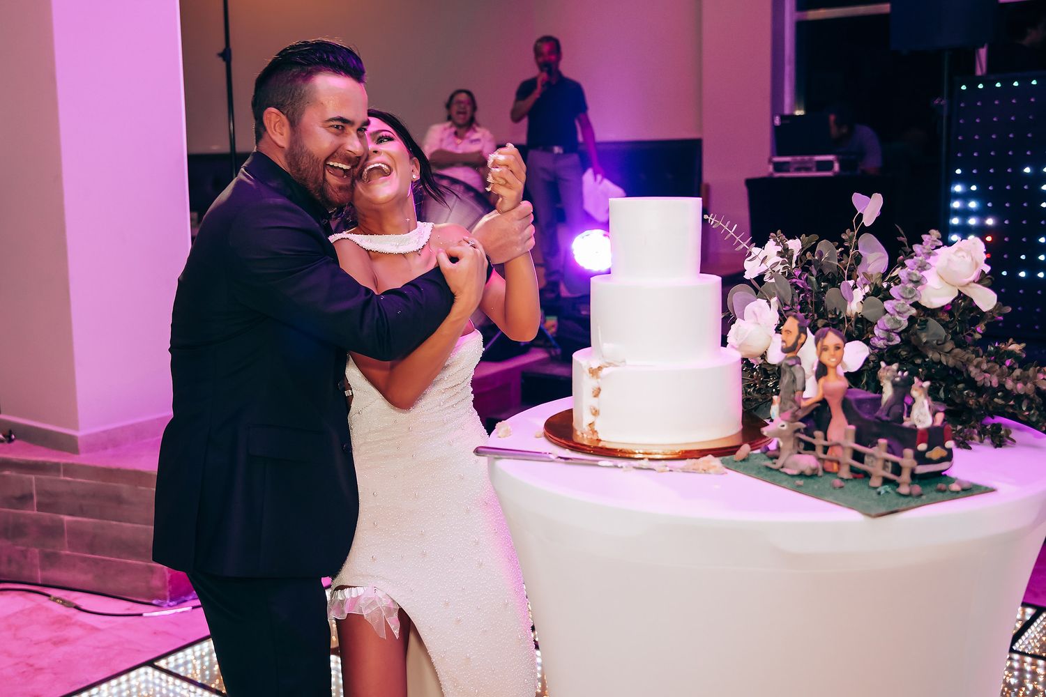 Wedding celebration with guests dancing and a white cake display under pink lighting.