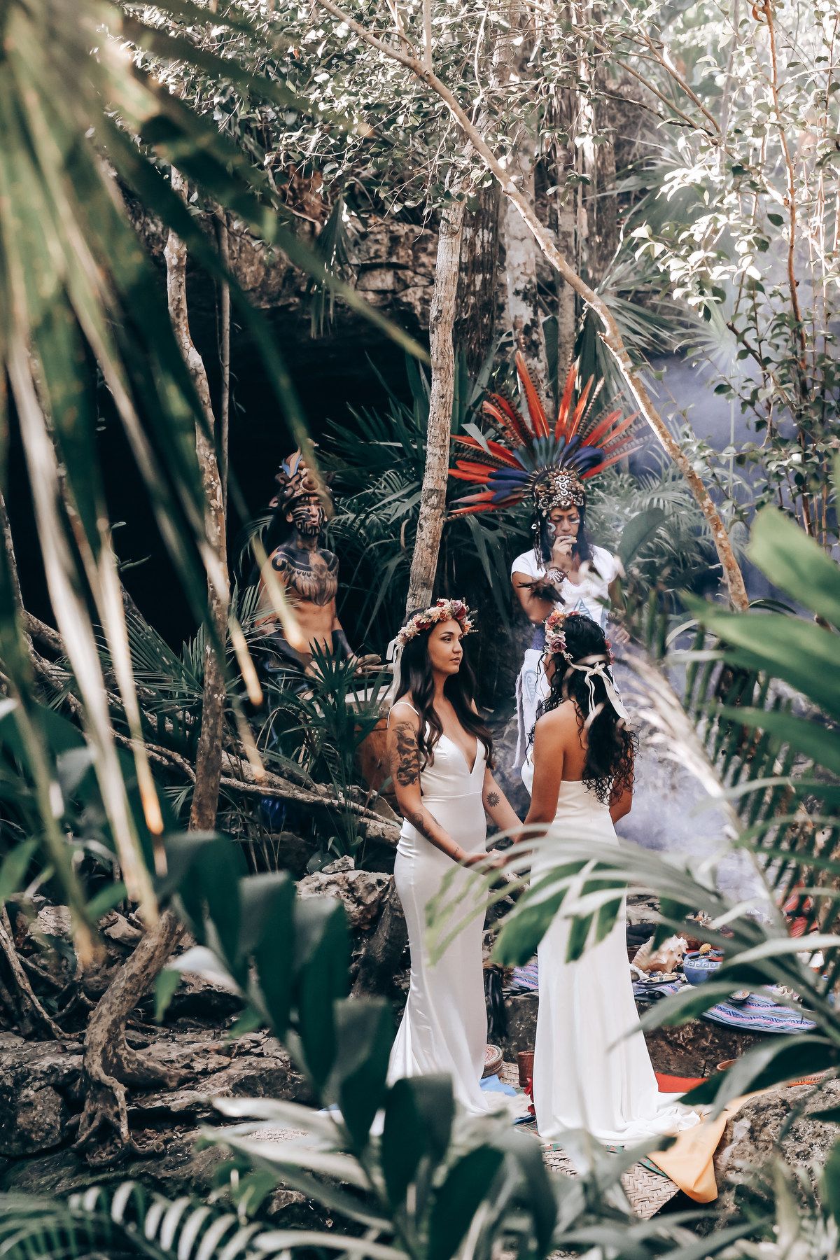 Two people in elegant white dresses walk through a lush tropical garden setting during a wedding photoshoot.