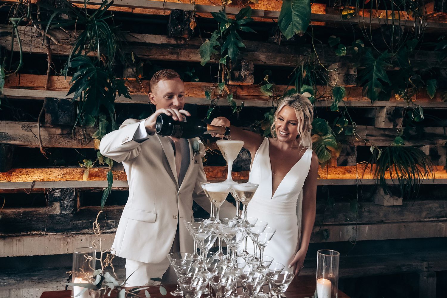Wedding party celebrates with raised glasses during an outdoor reception under wooden beams and hanging lights.
