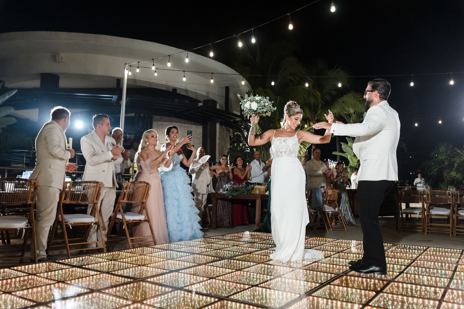 Bride and groom share first dance on illuminated floor at outdoor wedding reception with guests applauding at night.