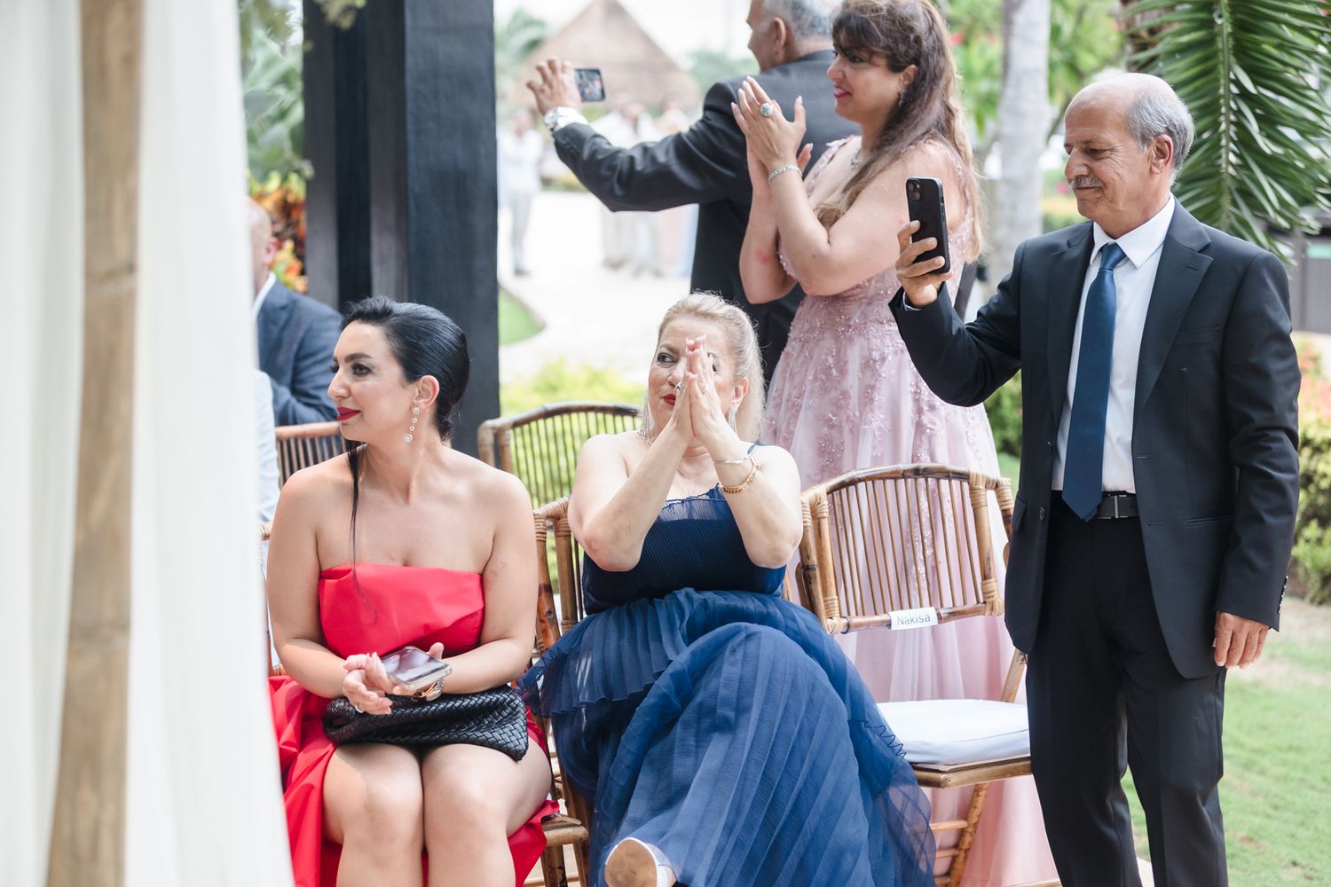Guests at an outdoor wedding ceremony, with women in elegant dresses reacting emotionally while others take photos.