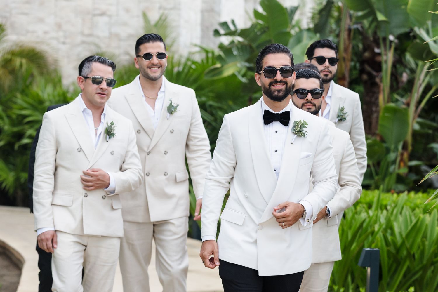 Groom in white tuxedo with bow tie leads groomsmen in matching white suits and sunglasses outdoors.