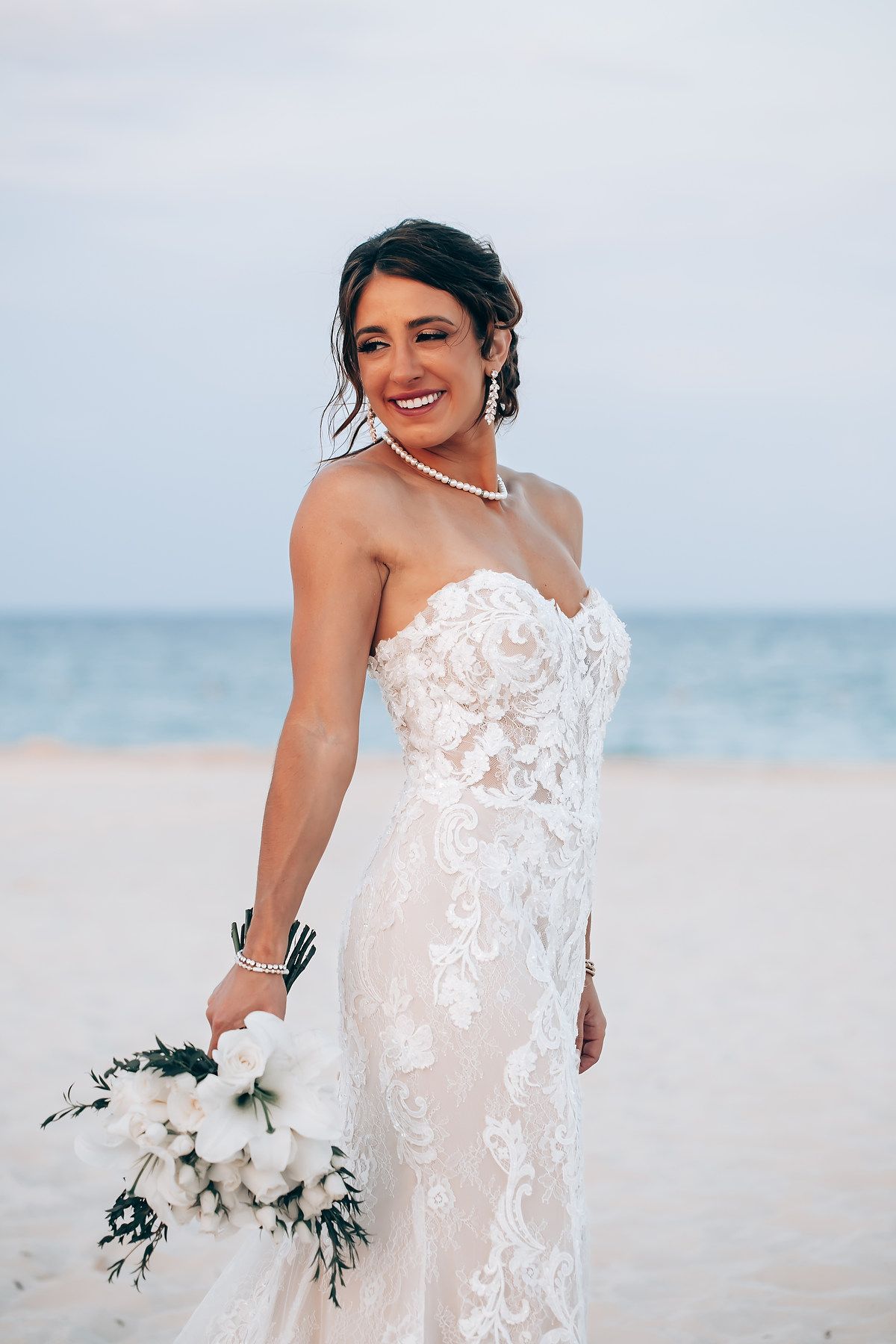 Bride in detailed lace gown holds white floral bouquet on scenic beach.