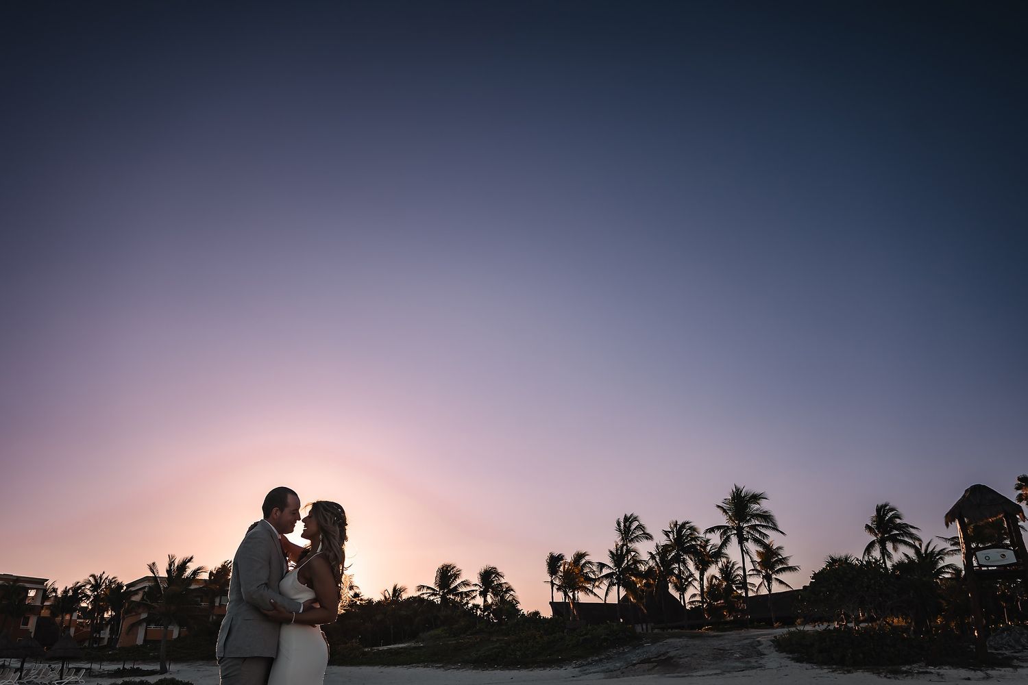 Silhouettes of palm trees against a dusky purple sunset sky at a beach wedding.