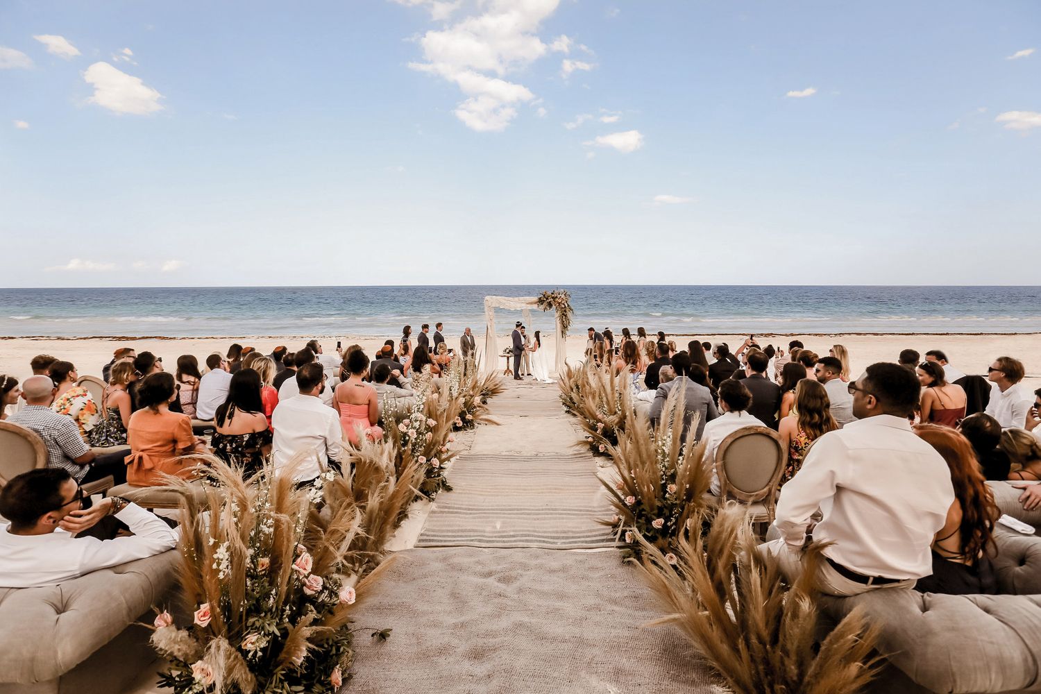 Panoramic view of a beach wedding ceremony setup with dried floral arrangements and guests.